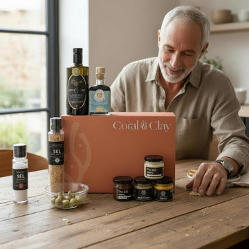 Lifestyle image featuring Coral & Clay For Dad gift box on a rustic wooden table, surrounded by gourmet items including olive oil, balsamic vinegar, seasoning blends, and jars of specialty salts, styled in a bright kitchen setting with natural light.