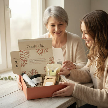 Lifestyle image featuring Coral & Clay For Mum gift box being given to mother on a light wooden table, filled with self-care items including a body lotion, bath salts, and a natural sponge, styled in a bright, cozy setting near a window.