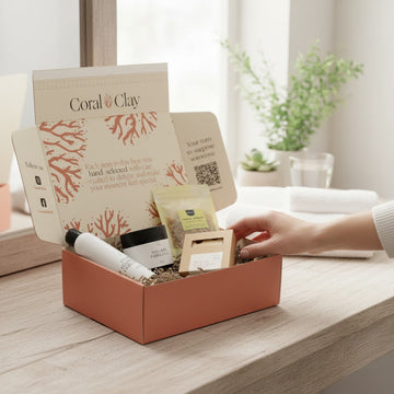 Coral & Clay signature gift box on a light wooden vanity, filled with self-care items including a body lotion, a black jar, a natural sponge, and a green-packaged product, styled with a folded white towel and a potted succulent.