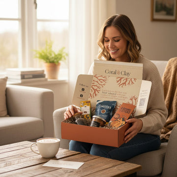 Lifestyle image featuring Coral & Clay anniversary gift box held on a wooden coffee table, filled with premium treats including artisan snacks, chocolate, and gourmet items, with a latte and card in a cozy living room setting with natural light.