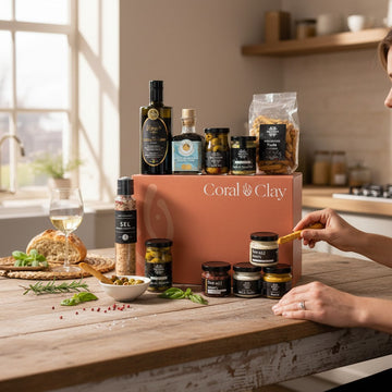 Lifestyle image featuring Coral & Clay housewarming gift box on a rustic wooden kitchen table, surrounded by gourmet items including olive oil, tapenade, crackers, and seasoning blends, with fresh bread and a glass of white wine in a cozy kitchen setting.