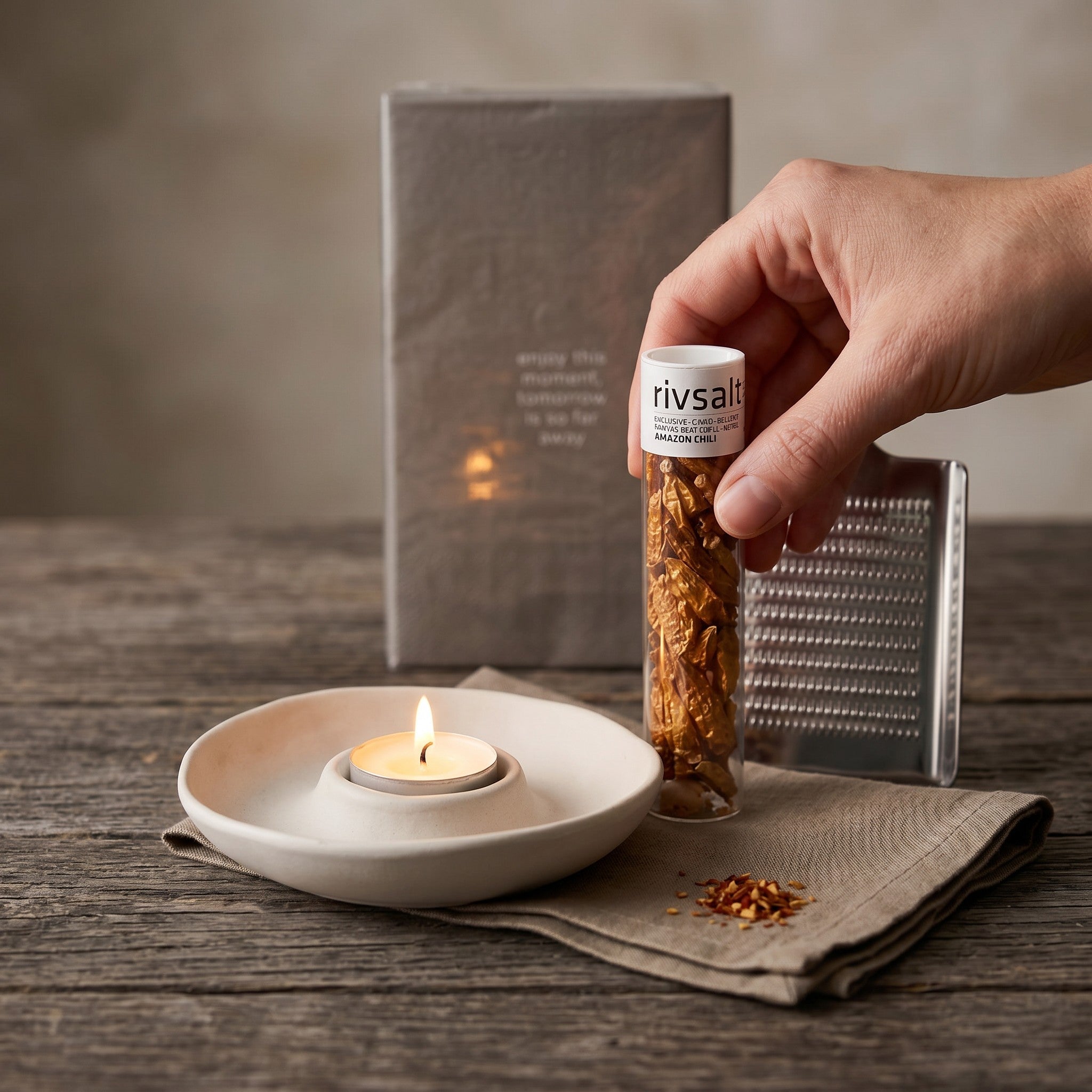 Hand holding a jar of 'rivsalt' next to a lit candle and grater on a wooden surface.