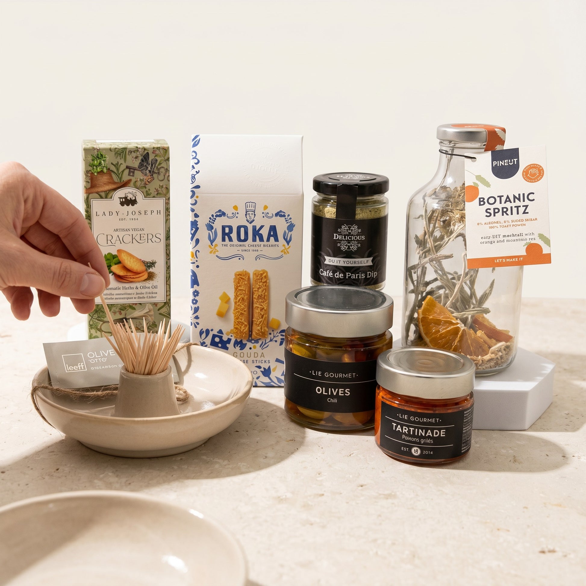 Aperitivo Moments Signature Gift assortment on white background: mocktail mix, chili olives, red pepper tapenade, dip mix, herb crackers, Gouda cheese sticks, olive bowl and napkins.