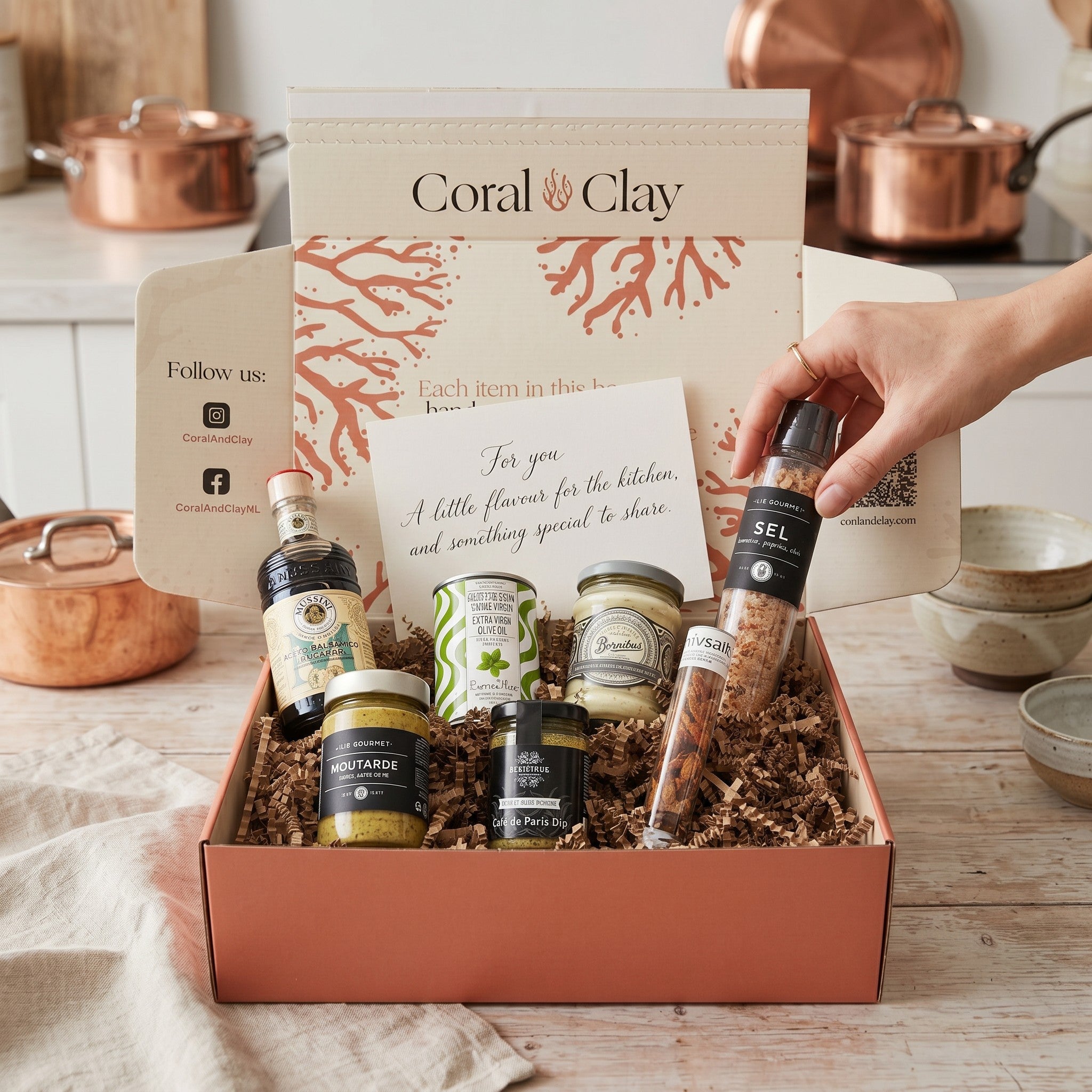 Gift box with various items including bottles and jars, with a hand reaching in to take one, set against a kitchen background.