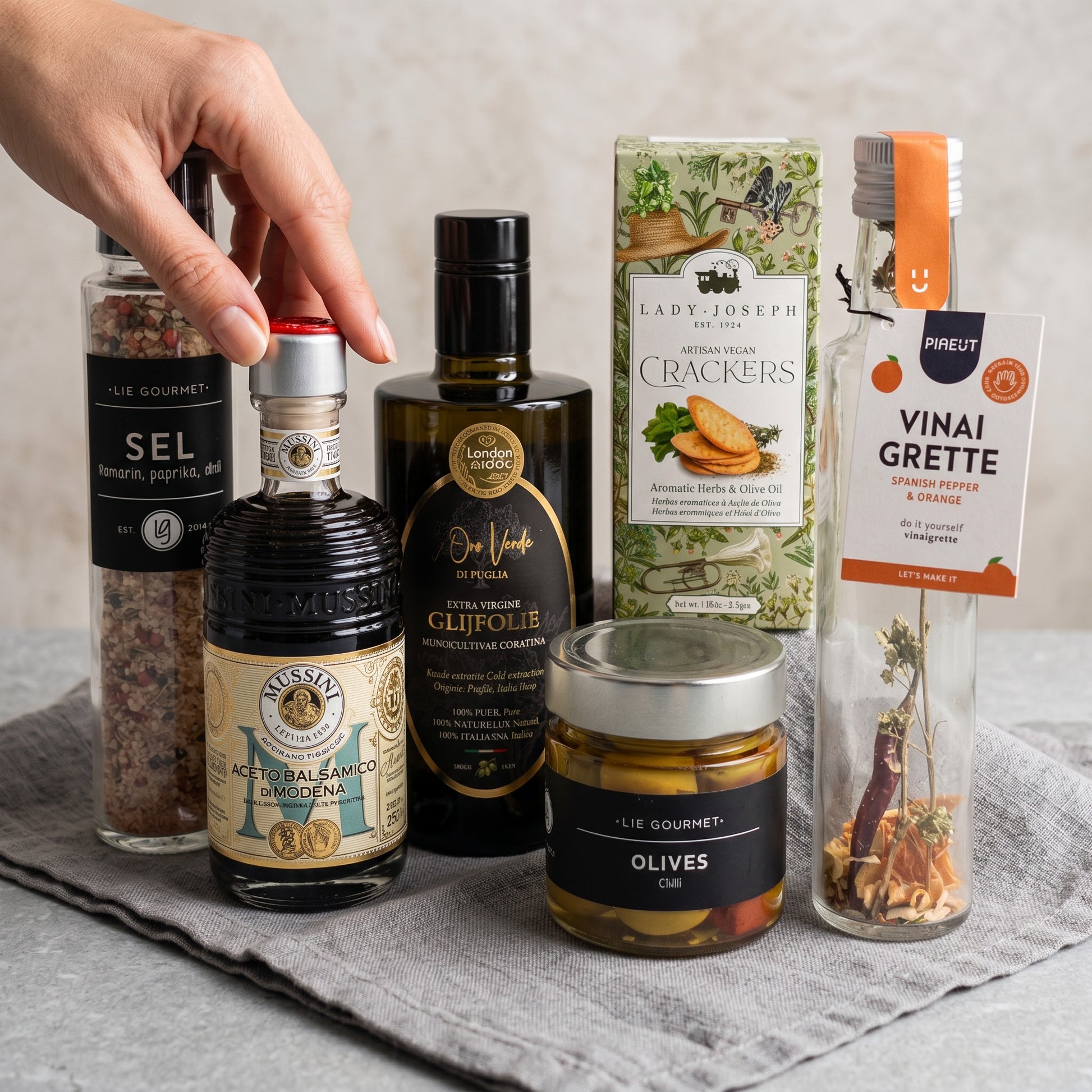 Luxury Mediterranean Signature Gift products on a gray surface: olive oil, balsamic vinegar, olives, crackers, vinaigrette and seasoning grinder.