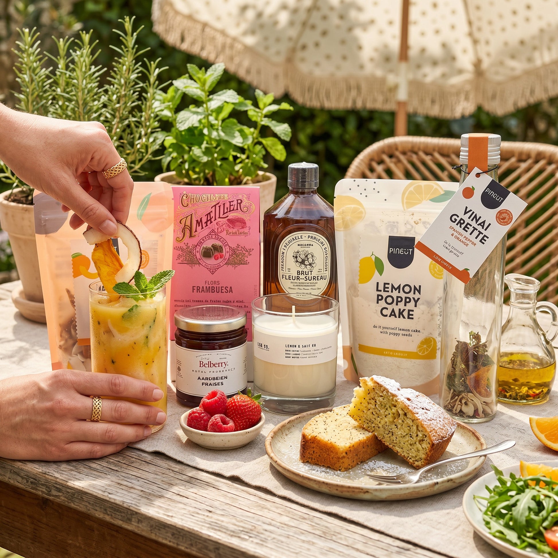 Coral & Clay Summer Feeling Signature Gift outdoor lifestyle photo – table scene with Mango Colada drink mix, elderflower syrup, lemon & poppy cake mix, strawberry confiture, raspberry pralines, Spanish pepper & orange vinaigrette, and scented candle.