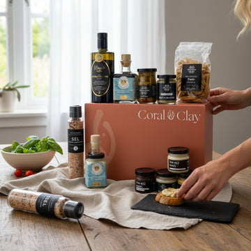 Lifestyle scene showcasing the Coral & Clay Grand Gourmet Gift Box on a wooden kitchen table. Premium deli items—olive oil, balsamic vinegar, pestos, olive mix, spice grinder, and breadsticks—are arranged around the box while someone prepares a slice of toasted bread with a gourmet spread. Fresh basil, tomatoes, and soft natural light complete the rustic cooking atmosphere.
