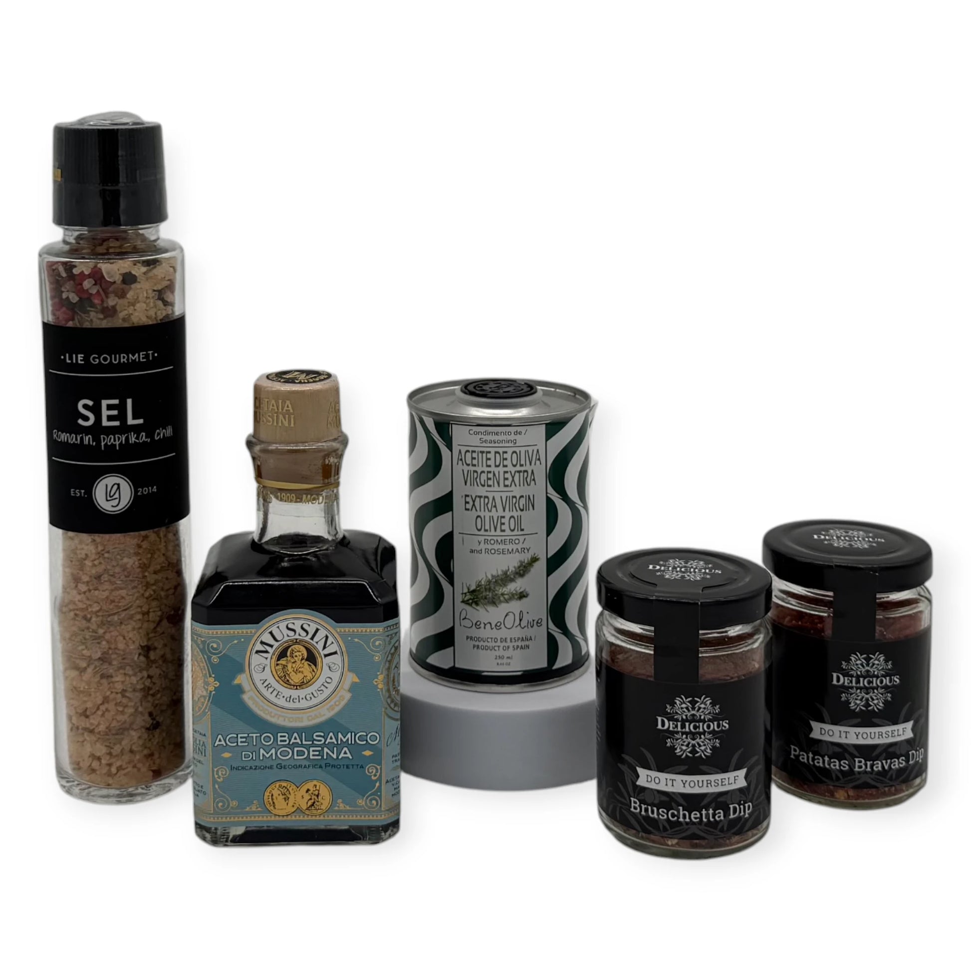 Five gourmet cooking items arranged on a white background: Lie Gourmet rosemary–paprika–chili salt grinder, Mussini Aceto Balsamico di Modena bottle, Beneoliva rosemary and oregano extra-virgin olive oil tin, Delicious Bruschetta dip jar, and Delicious Patatas Bravas dip jar.