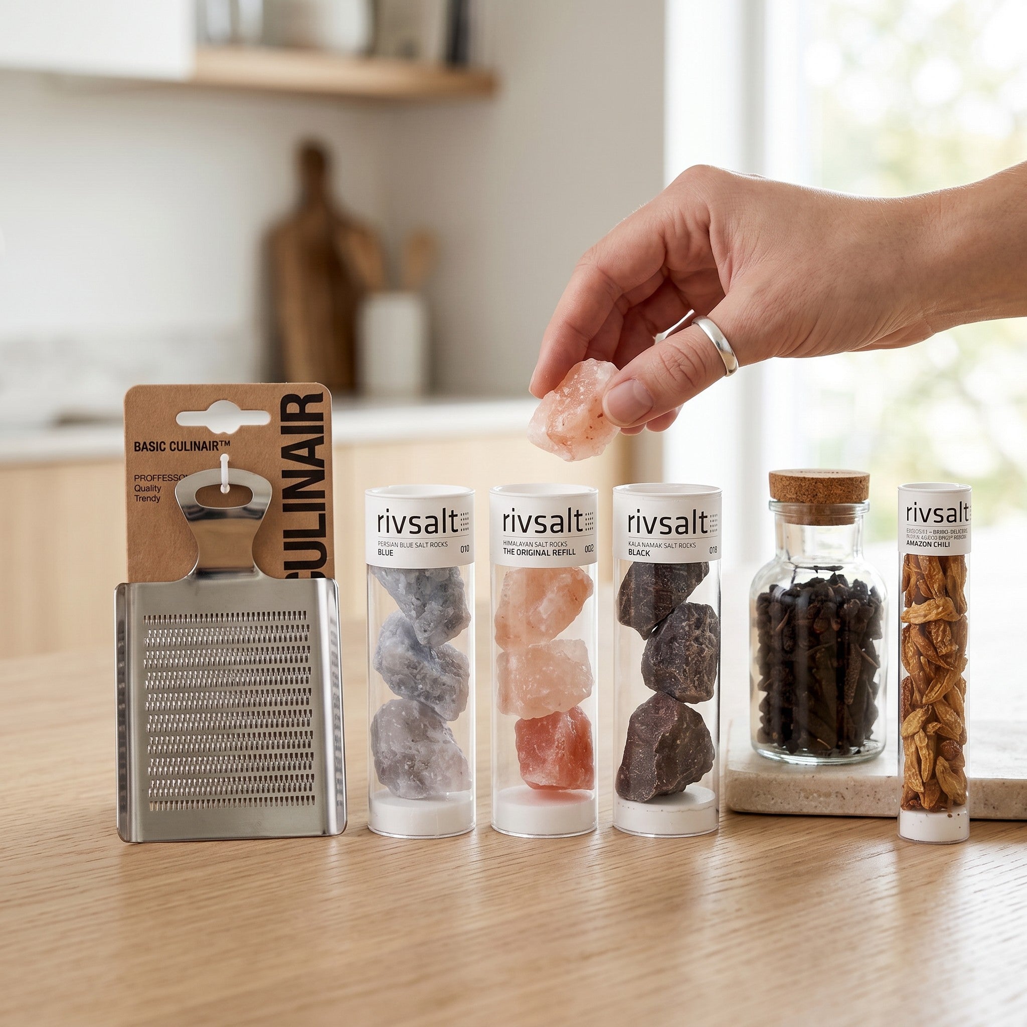 Coral & Clay Spiceworks Signature Gift lifestyle photo – hand reaching for Rivsalt gourmet salt on a kitchen counter with a stainless-steel grater and other salts and spices.