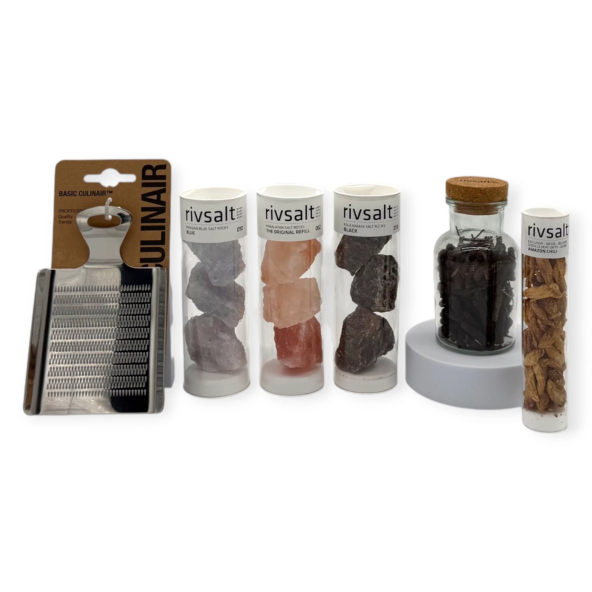 Rivsalt gourmet salt rock collection with grater set — including Persian blue salt rocks, Himalayan pink salt rocks, black Kala Namak salt rocks, Amazon chili refill tube, and a stainless-steel grater alongside a glass jar of whole spices.