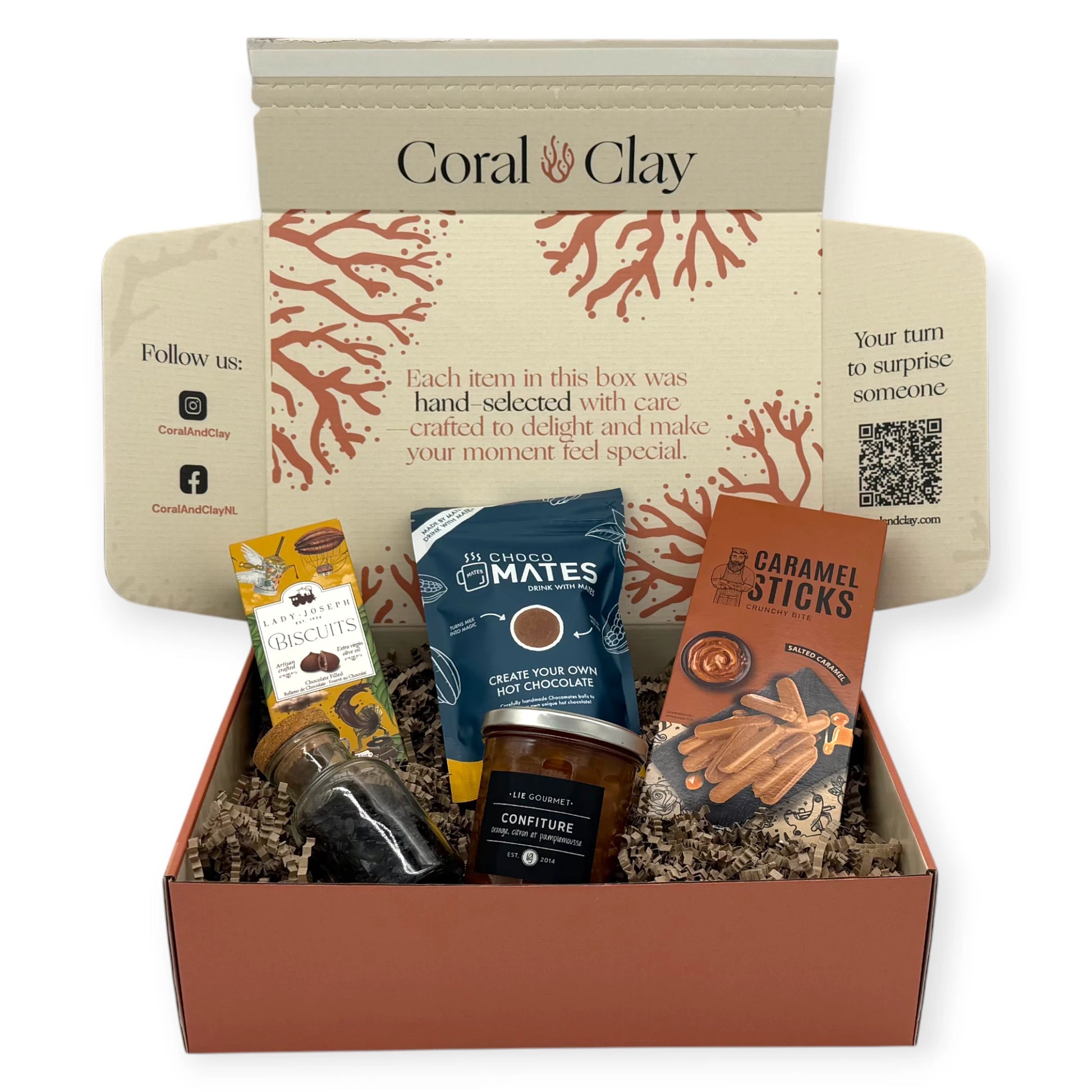 A top-down view of the Sunday Afternoon Signature Gift inside the Coral & Clay box, showing caramel sticks, hot chocolate mix, chocolate biscuits, fruit confiture, and a small bottle of chocolate flakes nestled in kraft paper filling.