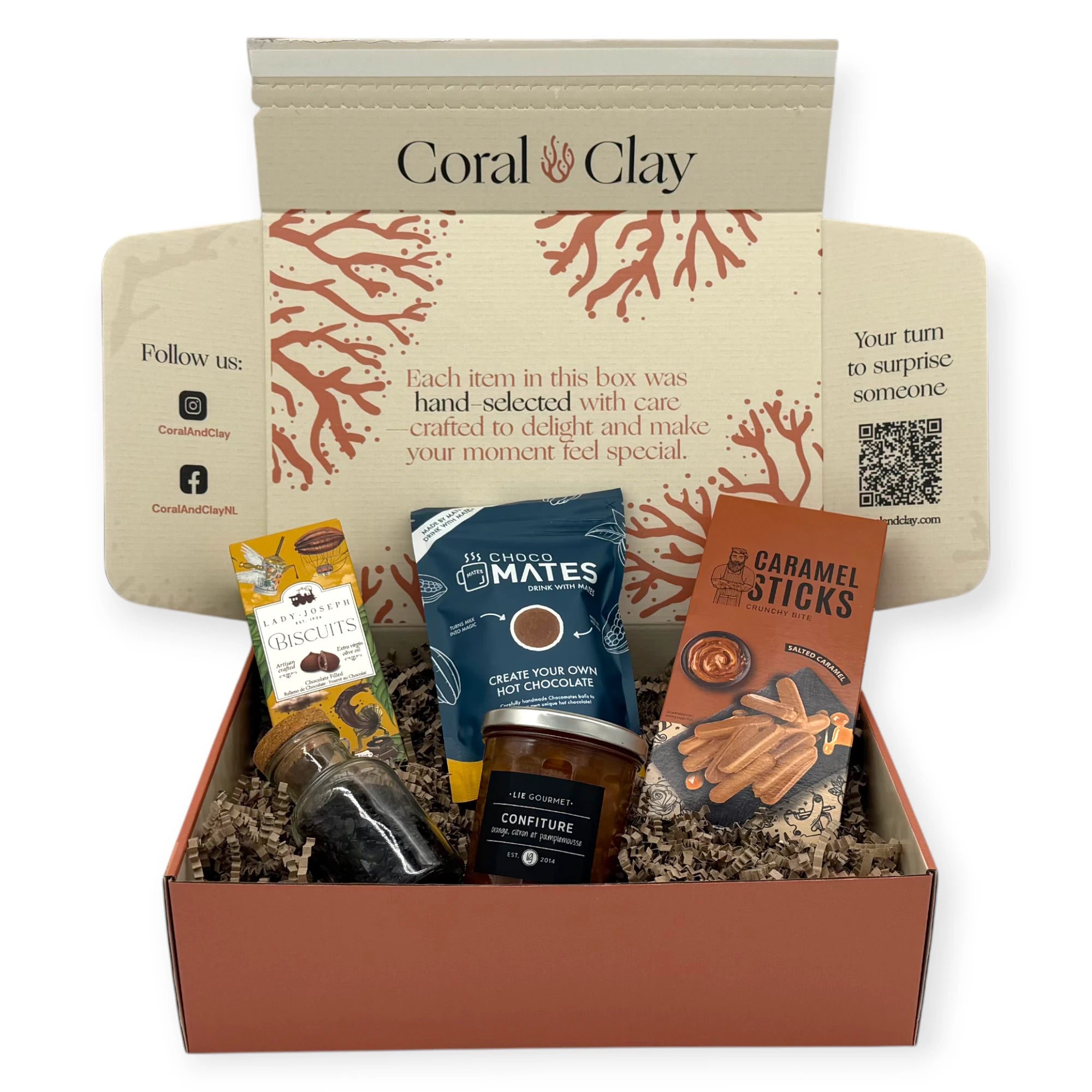 A top-down view of the Sunday Afternoon Signature Gift inside the Coral & Clay box, showing caramel sticks, hot chocolate mix, chocolate biscuits, fruit confiture, and a small bottle of chocolate flakes nestled in kraft paper filling.